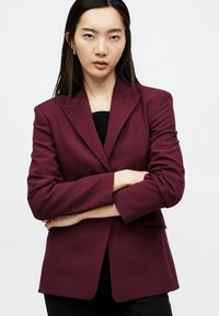 Νεαρή γυναίκα με ίσια σκούρα μαλλιά, φοράει μπορντό σακάκι και μαύρο τοπ, με σταυρωμένα χέρια και κοιτάζει με αυτοπεποίθηση την κάμερα.