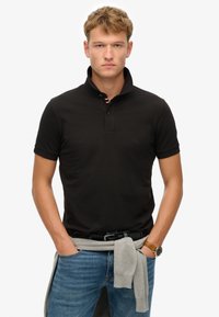 Polo shirt nera con maniche corte, abbottonatura e dettagli del colletto a righe. Indossata con jeans blu e un maglione grigio legato in vita.