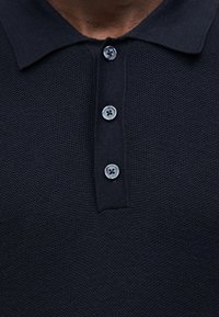 Polo bleu marine avec un motif tricoté texturé, doté d'un col et de trois boutons gris au niveau du cou. Gros plan sur le détail du tissage du tissu.