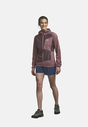 Frau lächelnd, trägt eine violette Kapuzenjacke, marineblaue Shorts und Trailrunning-Schuhe, steht mit leicht gespreizten Beinen vor weißem Hintergrund.