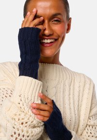 Personne souriante portant un pull crème en tricot torsadé et des gants sans doigts bleu marine, avec une main couvrant partiellement son visage.