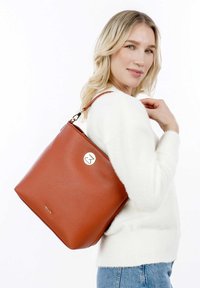 Mujer rubia con suéter blanco y jeans lleva un gran bolso de piel marrón con detalles dorados, mirando por encima del hombro.