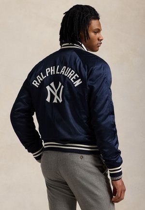 Νεαρός άντρας φοράει μπλε ναυτικό μπουφάν Ralph Lauren με λευκές ρίγες στις μανσέτες και τη μέση, γκρι παντελόνι, γυρισμένος πλαγίως δείχνοντας το λογότυπο στην πλάτη.