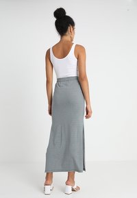 Femme aux cheveux bouclés relevés en chignon, portant un haut sans manches blanc, une jupe maxi à rayures taille haute avec une fente sur le côté, et des sandales à talons blocs blanches.