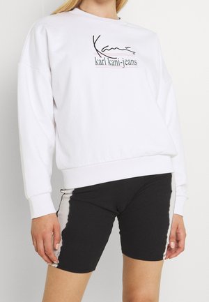 Hvid sweatshirt med sort "karl kani-jeans"-logo kombineret med sorte og hvide tie-dye cykelshorts på en stående person.