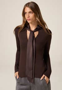 Maglietta marrone a maniche lunghe con scollo a chiave e cravatta in satin attaccata. Materiale morbido e aderente con cuciture pronunciate, abbinata a pantaloni grigi a righe.