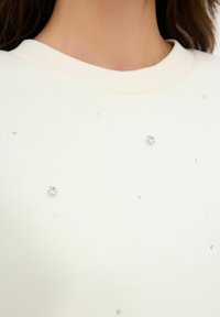 Bílá mikina se širokým výstřihem, dekórávaná roztroušenými kamínky různých velikostí pro zvýšení detailu a lesku. Měkká textura.