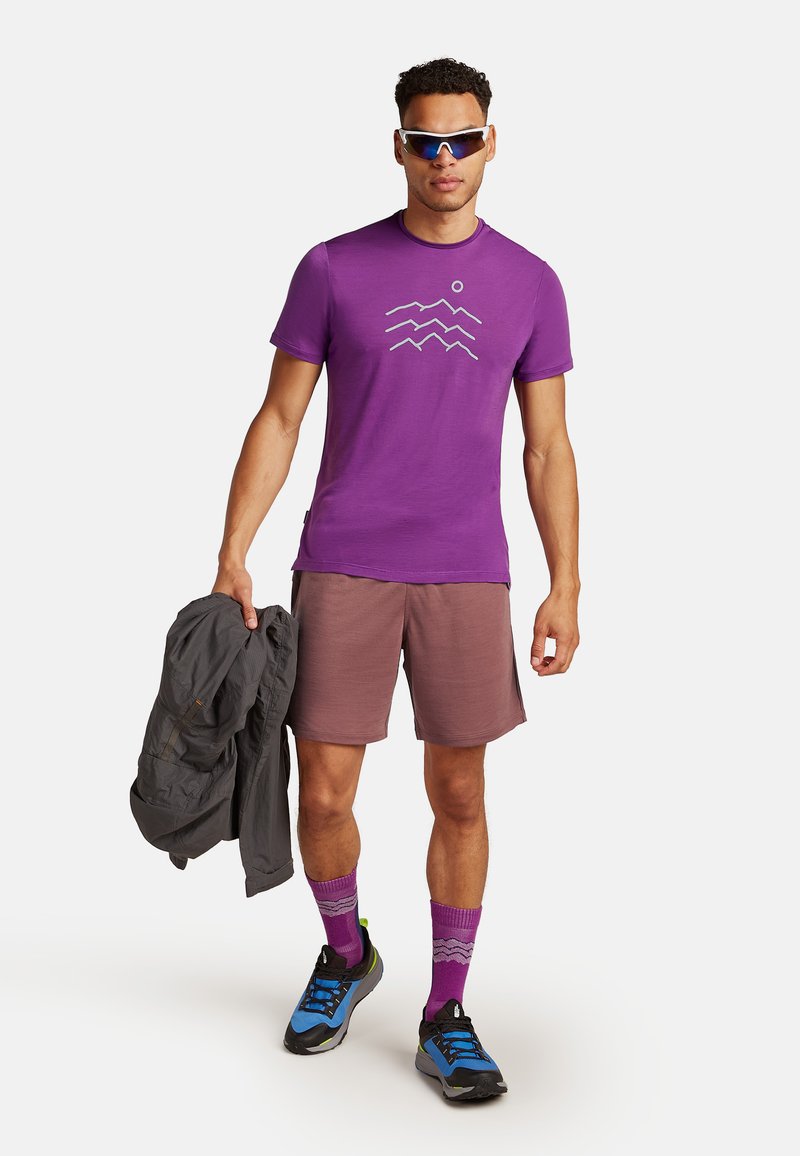 Paarse sport T-shirt met golfpatroon, gecombineerd met lichtbruine shorts, blauwe en zwarte schoenen en paarse sokken. Grijze jas in de hand.