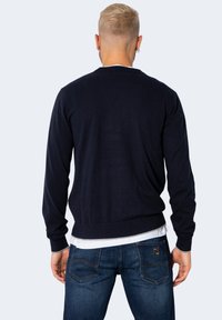 Maglione lavorato a maglia blu navy con maniche lunghe, polsini e orlo a costine. Maglietta bianca visibile al colletto, abbinata a jeans blu.