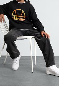 En person sitter på en vit stol iförd en svart ellesse-tröja, mörka manchesterbyxor och vita Nike-sneakers på ett grått golv.