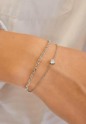 Due delicati braccialetti d'argento al polso, uno con un piccolo ciondolo a forma di cuore.