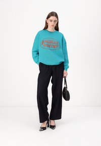 Sweatshirt oversized em verde água com a estampa "AMERICAN VINTAGE", combinado com calças largas de riscas finas em preto e saltos altos pretos, segurando uma bolsa preta.