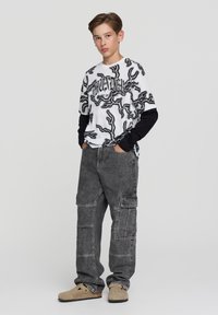 Nastolatek stojący z rękami w kieszeniach, ubrany w czarno-białą koszulkę z grafiką, szare spodnie cargo i beżowe buty wsuwane na tle białego tła.