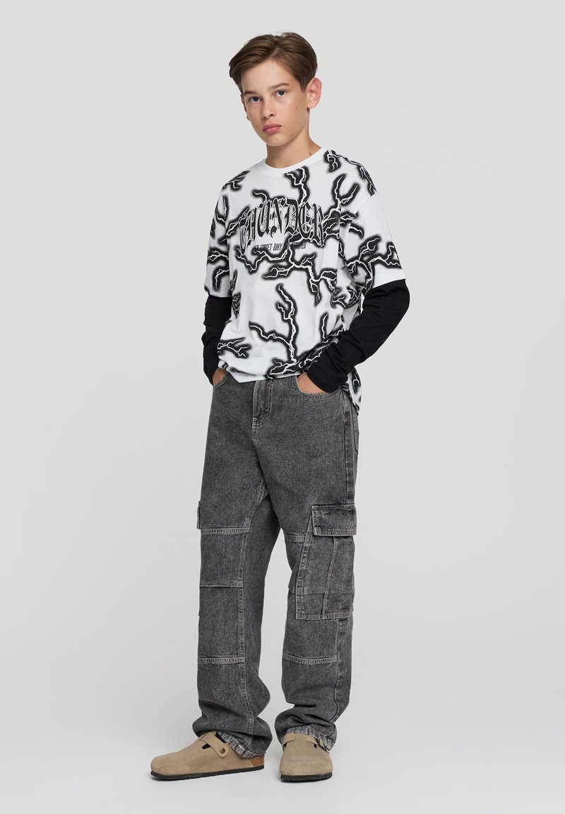 Nastolatek stojący z rękami w kieszeniach, ubrany w czarno-białą koszulkę z grafiką, szare spodnie cargo i beżowe buty wsuwane na tle białego tła.