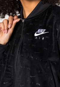 Närbild av en persons hand som öppnar dragkedjan på en svart Nike Air-jacka med broderade logotyper och strukturerade mönster.