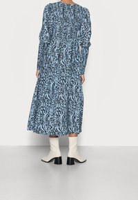 Robe longue bleue avec un imprimé abstrait noir, dotée de manches bouffantes et d'un design froncé. Stylisée avec des bottines blanches et un ourlet évasé.