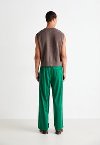 Suéter marrón sin mangas con un dobladillo acanalado, combinado con pantalones verdes de corte holgado. El conjunto presenta texturas suaves y un corte contemporáneo.