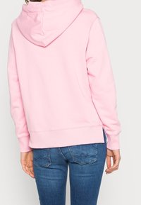 Persona con sudadera con capucha rosa claro y jeans azules, vista desde atrás sobre un fondo liso.