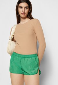 Calções verdes, leves, com cintura elástica e fendas laterais, combinados com uma blusa de mangas longas em bege com nervuras. Logótipo de texto notável nos calções.