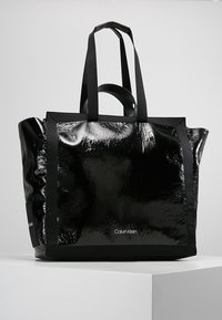 Czarna torba na ramię o błyszczącej, teksturowanej powierzchni. Wzmocnione uchwyty i kontrastowe czarne wykończenie. Na boku znajduje się logo Calvin Klein.
