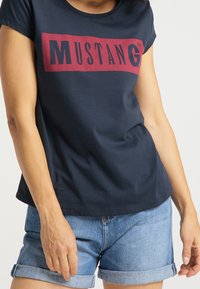 Dark blue cotton t-shirt with short sleeves, featuring a red "MUSTANG" logo printed across the chest. Paired with light denim shorts.