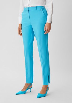 Vrouw draagt aansluitende fel turquoise broek met voorplooi, zijsplit aan zoom, gecombineerd met bijpassende puntige slingback hakken en wit overhemd.