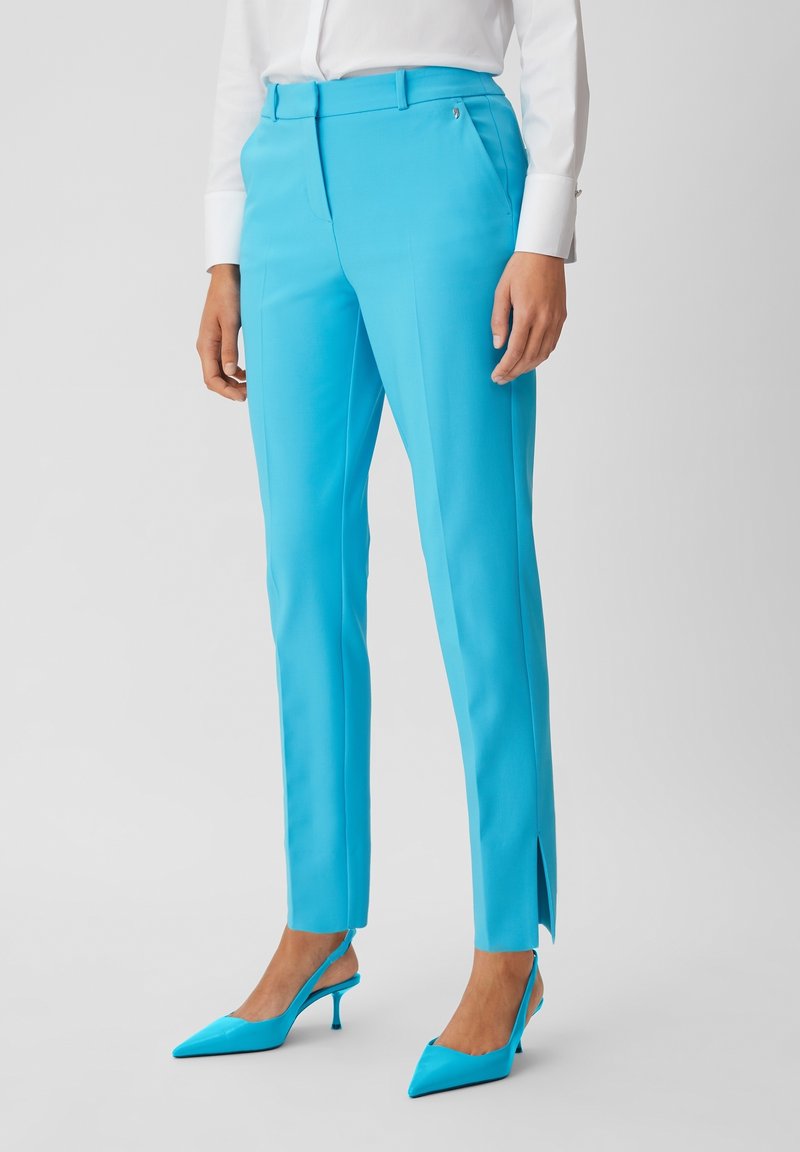 Femme portant un pantalon turquoise vif ajusté avec pli sur le devant, ourlet fendu sur le côté, associé à des talons slingback pointus assortis et une chemise blanche.