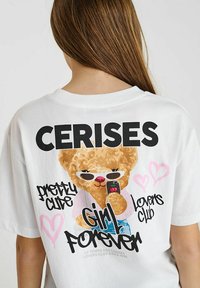 T-shirt blanc avec un ours en cartoon portant des lunettes de soleil et tenant un téléphone. Le texte inclut "CERISES," "plutôt mignon," "Fille pour toujours," et des cœurs.