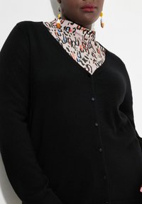 Svart cardigan med V-ringning, med svarta knappar, dragen över en mönstrad polotröja i beige, svart, röd och blå.