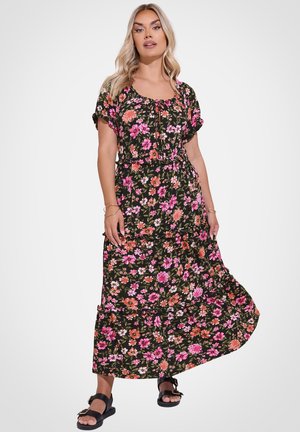 Donna con lunghi capelli biondi che indossa un abito lungo nero a fiori e sandali neri con fibbia, in piedi davanti a uno sfondo bianco.