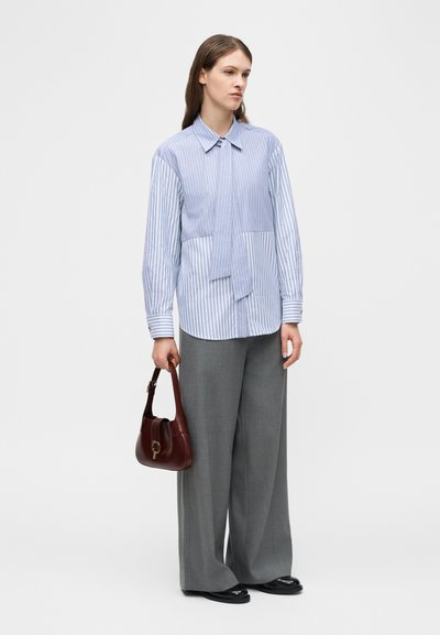 Γυναίκα με μπλούζα μακρυμάνικη ριγέ σε δύο αποχρώσεις του μπλε, φαρδιά γκρι παντελόνια, μαύρα παπούτσια και κρατώντας καφέ σκούρη τσάντα χειρός.