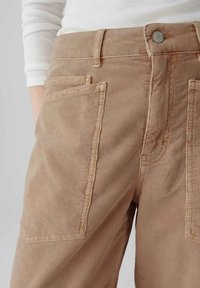 Pantalons en velours côtelé beige avec un tissu texturé, de larges poches avant et des détails de couture visibles. Bouton en métal à la taille.
