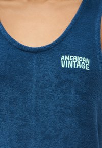 Κοντινό πλάνο ενός ατόμου που φοράει μπλε ριγωτό αμάνικο μπλουζάκι με το κέντημα «American Vintage» σε λευκό στο στήθος.