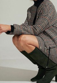 Femme accroupie portant un ensemble manteau et jupe à motif pied-de-poule avec un col roulé noir et des bottes hautes en daim vert foncé.