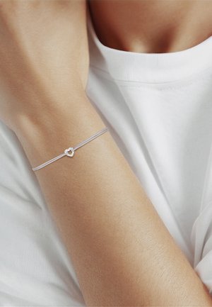 Delicada pulsera de plata con un pequeño colgante en forma de corazón en una muñeca, llevada con una camiseta blanca de manga corta.