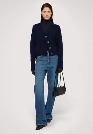 Cárdigan de punto azul marino con escote en V y tres botones grandes, combinado con jeans acampanados azules y guantes negros, sosteniendo un pequeño bolso negro.