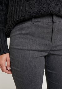 Kolsvarta byxor i en slät, texturerad tyg. Har en framvik, bälteshällor och en smal passform. Hand delvis synlig.