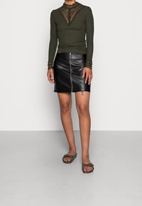 Olivgrön långärmad topp med spetsdetaljer, kombinerad med en svart läderkortkjol med dragkedja och olivgröna sandaler.