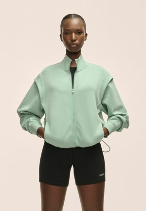 Sudadera de color verde menta con cremallera, hombros caídos, tela texturizada y bolsillos laterales. Se lleva con pantalones cortos negros que presentan un logo.