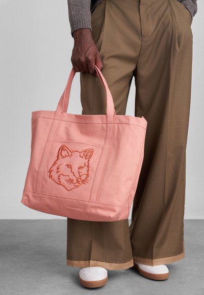 Ροζ καμβάς τσάντα tote με κεντητό σχέδιο αλεπού. Διαθέτει ανθεκτικά χερούλια και ορθογώνιο σχήμα, φορεμένη από ένα άτομο με μπεζ παντελόνια.