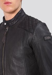 Giacca di pelle nera con design trapuntato sulle spalle, chiusura con cerniera e dettagli in metallo. Presenta un colletto alto e una superficie strutturata.