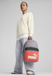 Sivý a oranžový batoh s logom Puma, s okrúhlym dizajnom a čiernou hornou časťou. Model nosí svetložltý sveter a voľné džínsy.