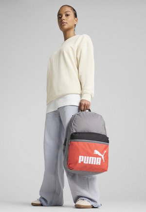 Grå og orange rygsæk med et Puma-logo, der har et rundt design og en sort topsektion. Modellen bærer en lys cremefarvet sweatshirt og løse jeans.