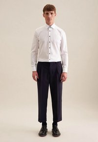 Camicia bianca con bottoni neri, abbinata a pantaloni scuri plissettati. Scarpe nere completano l'outfit, creando un aspetto formale.