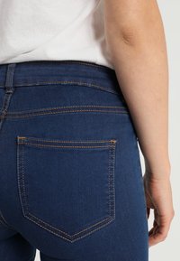 Jeans de mezclilla azul oscuro con un corte ajustado, que cuentan con un único bolsillo trasero, costuras visibles en un color naranja contrastante y una textura suave.