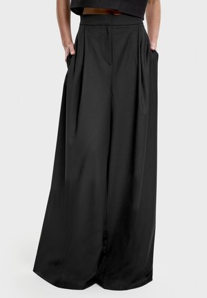 Osoba ubrana w czarne, wysokie spodnie o szerokich nogawkach, z rękami w kieszeniach na jednolitym szarym tle.