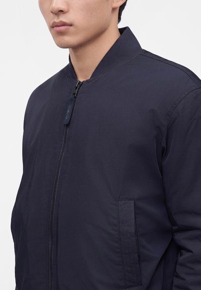 Gros plan d'une personne portant une veste bomber bleu marine foncé avec une fermeture éclair et un col côtelé visible sur un fond uni.