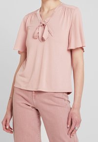 Różowa bluzka z krótkim rękawem z miękkiego materiału, z dekoltem w kształcie litery V i wiązaniem z przodu, z delikatnym marszczeniem na ramionach.
