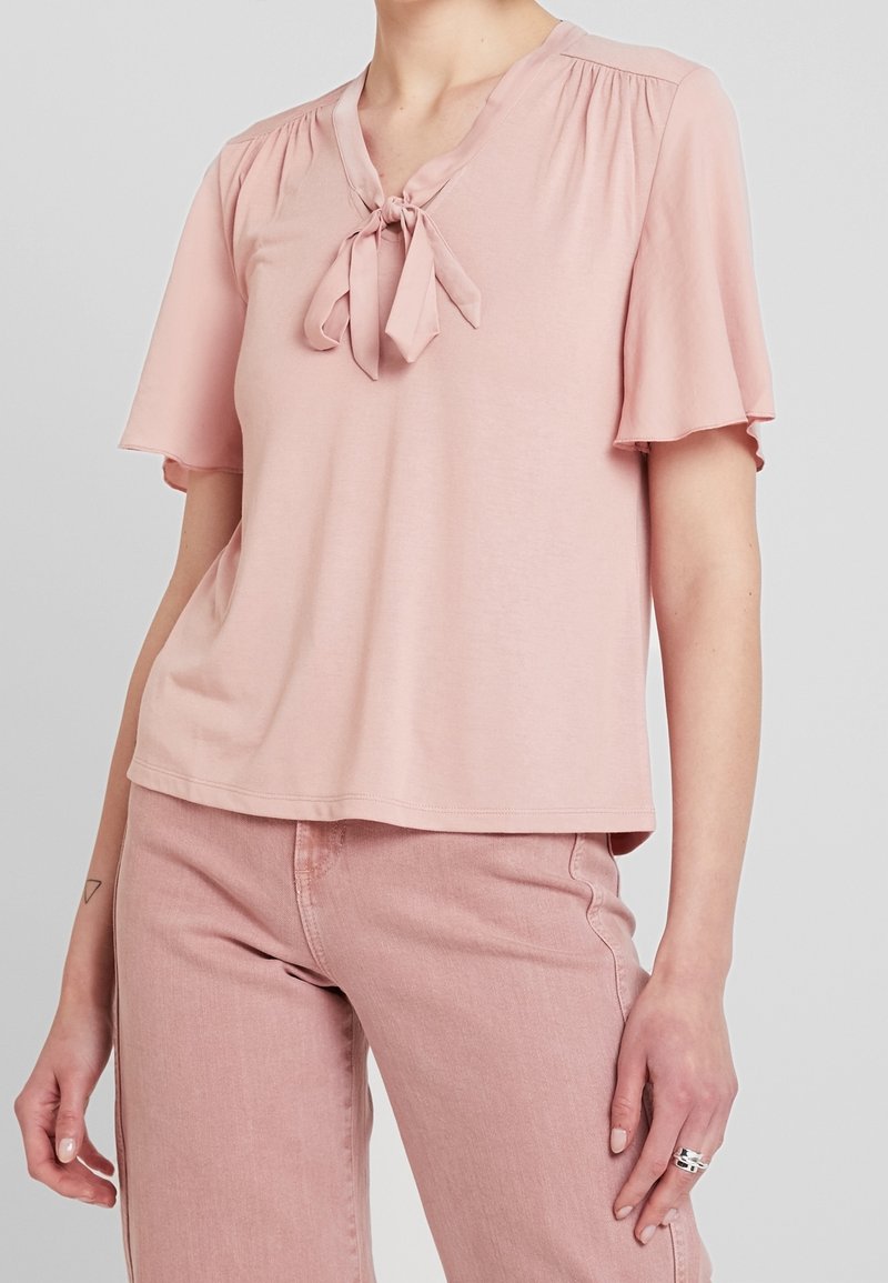 Różowa bluzka z krótkim rękawem z miękkiego materiału, z dekoltem w kształcie litery V i wiązaniem z przodu, z delikatnym marszczeniem na ramionach.