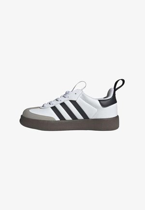 Zapatilla deportiva blanca con detalles en cuero negro, tres rayas negras, puntera de goma gris y suela de brown texturizada. Cordones en gris claro.